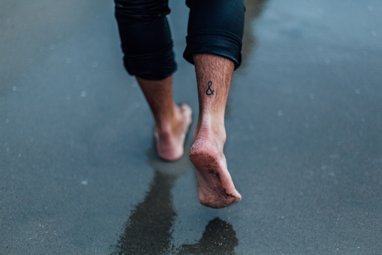 Die nackten Knöchel und Füße einer Person. Die Hose ist etwas hochgekrempelt. Ein Tattoo von einem kaufmännischen “und” wird sichtbar.