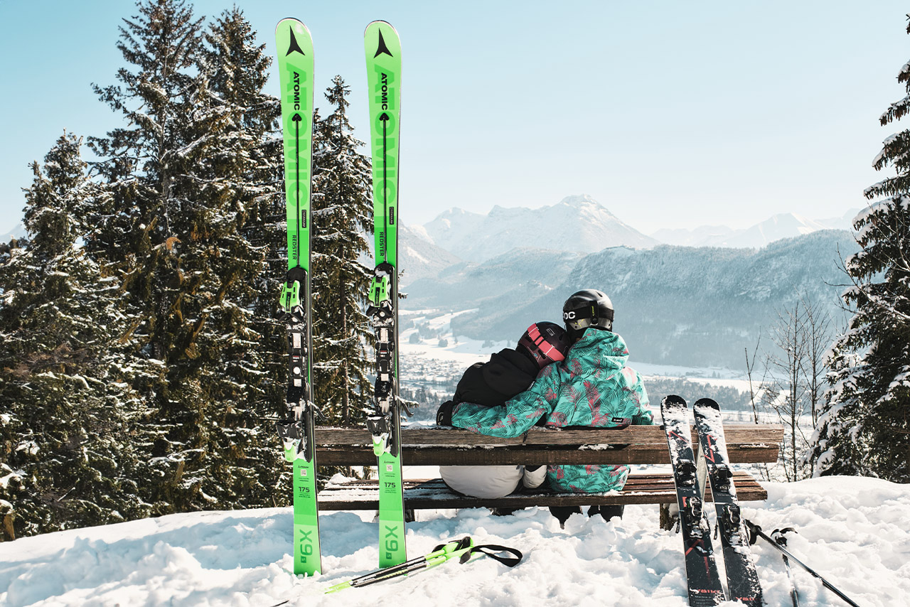 Ein Paar, das Arm in Arm auf einer Bank in der verschneiten Schneelandschaft sitzt. Ihre Skier lehnen an der Bank.